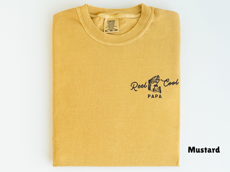 Op de afbeelding: Mosterdkleurig T-shirt met de tekst "Reel Cool Papa" en een visafbeelding. Het gevouwen shirt heeft een label met de tekst "Comfort Colors XL 10-20".