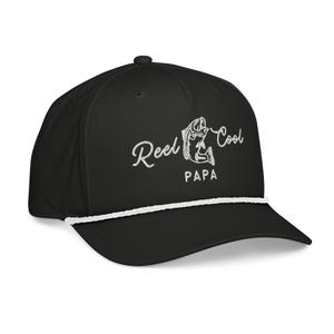 May include: Black baseball cap with a white rope detail and a curved brim. The cap features the embroidered text "Reel Cool Papa" in white script and a fish graphic. A stylish accessory for fishing enthusiasts.