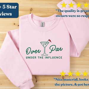 May include: Light pink sweatshirt with the text "Over Par UNDER THE INFLUENCE" in green, featuring a martini glass with a golf flag. The sweatshirt has long sleeves and includes customer reviews.