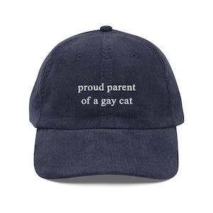 May include: A navy blue corduroy baseball cap with white embroidered text that reads "proud parent of a gay cat".