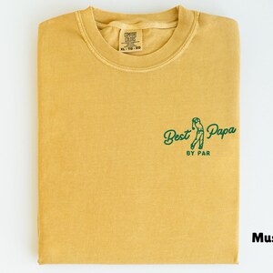 Op de afbeelding: Mosterdkleurig T-shirt met de tekst "Best Papa by Par" in groen. Het shirt is opgevouwen en heeft een ronde hals. Het label op het shirt luidt "Comfort Colors XL 10-20".