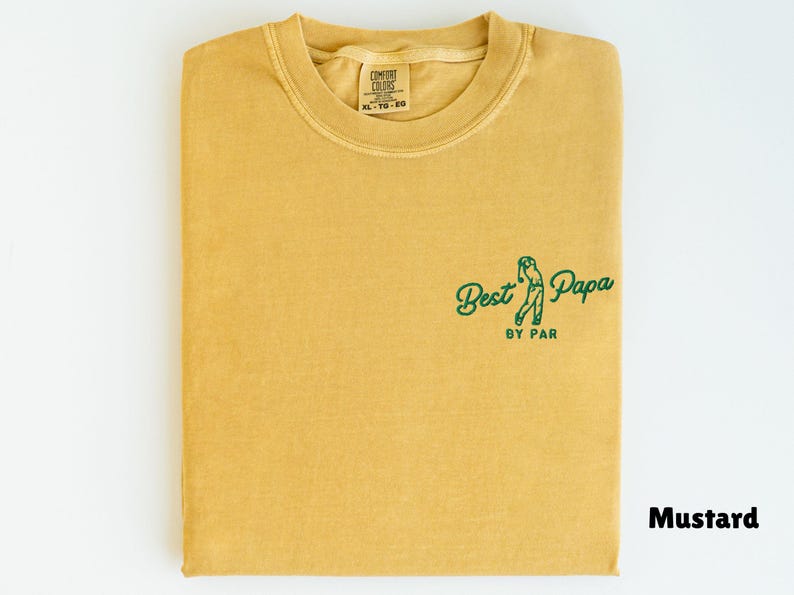 Op de afbeelding: Mosterdkleurig T-shirt met de tekst "Best Papa by Par" in groen. Het shirt is opgevouwen en heeft een ronde hals. Het label op het shirt luidt "Comfort Colors XL 10-20".