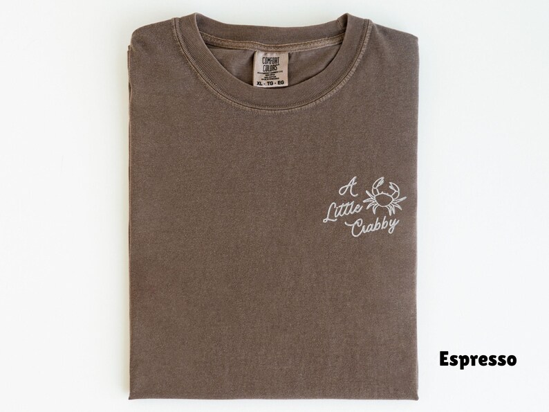 May include: Espresso-colored t-shirt with the phrase "A Little Crabby" and a crab graphic embroidered in white. The shirt is folded neatly, with the brand tag visible at the neckline. The color is a muted brown.