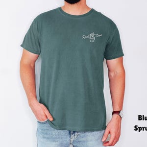 Pu&ograve; includere: T-shirt blu abete con la scritta "Reel Cool Dad" in corsivo bianco. La maglietta &egrave; a tinta unita e ha maniche corte. Il modello indossa jeans blu e un orologio.