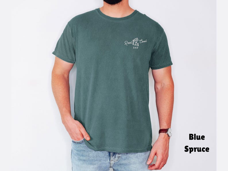 Pu&ograve; includere: T-shirt blu abete con la scritta "Reel Cool Dad" in corsivo bianco. La maglietta &egrave; a tinta unita e ha maniche corte. Il modello indossa jeans blu e un orologio.