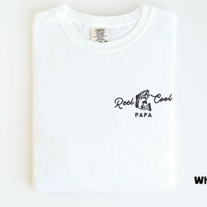 Op de afbeelding: Wit T-shirt met een ronde hals, netjes opgevouwen. Het shirt heeft een zwarte afbeelding van een vis en de woorden "Reel Cool Papa" in een scriptlettertype. Het shirt is gemaakt van een zacht materiaal.