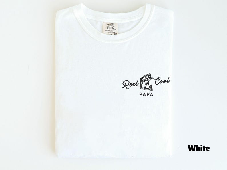 Op de afbeelding: Wit T-shirt met een ronde hals, netjes opgevouwen. Het shirt heeft een zwarte afbeelding van een vis en de woorden "Reel Cool Papa" in een scriptlettertype. Het shirt is gemaakt van een zacht materiaal.