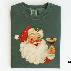 May include: A folded moss green t-shirt featuring a vintage illustration of Santa Claus holding a cocktail. Santa is wearing a red and white hat with holly berries. The shirt has the word "Moss" printed on the bottom right.