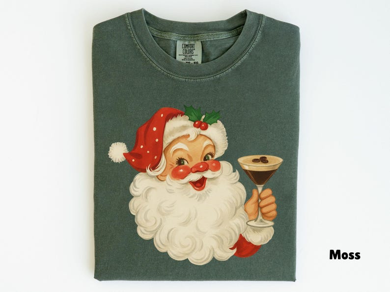 May include: A folded moss green t-shirt featuring a vintage illustration of Santa Claus holding a cocktail. Santa is wearing a red and white hat with holly berries. The shirt has the word "Moss" printed on the bottom right.