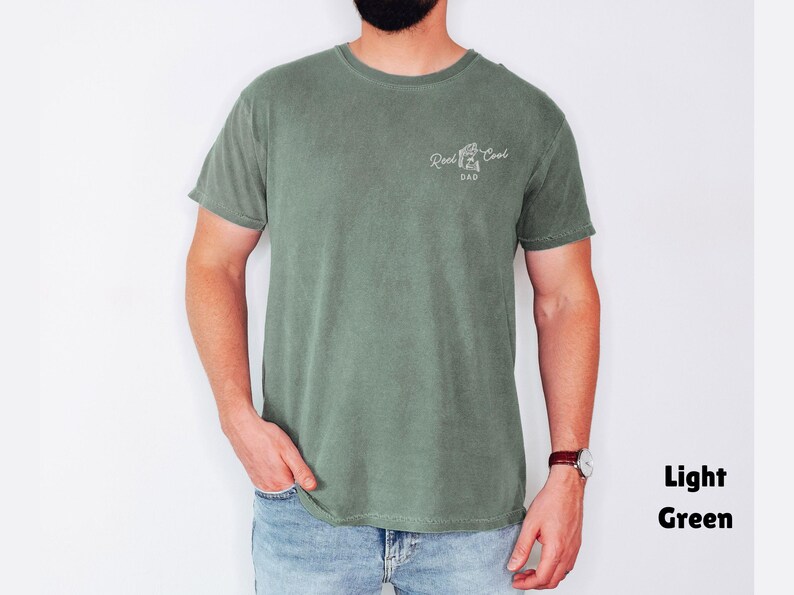 Pu&ograve; includere: T-shirt verde chiaro con la scritta bianca "Reel Cool Dad". La maglietta &egrave; tinta unita e ha lo scollo tondo. Il modello indossa jeans blu e un orologio.