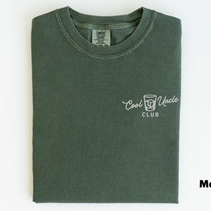 May include: A moss green t-shirt with the words "Cool Uncle Club" and a graphic of a cocktail glass printed in white. The shirt is folded neatly on a white surface.