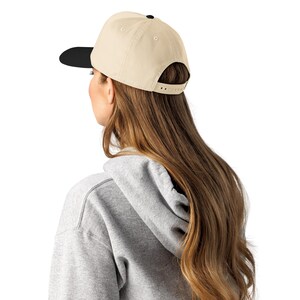 May include: A beige baseball cap with a black brim and a black button on top. The cap is worn by a person with long brown hair and a gray hoodie.