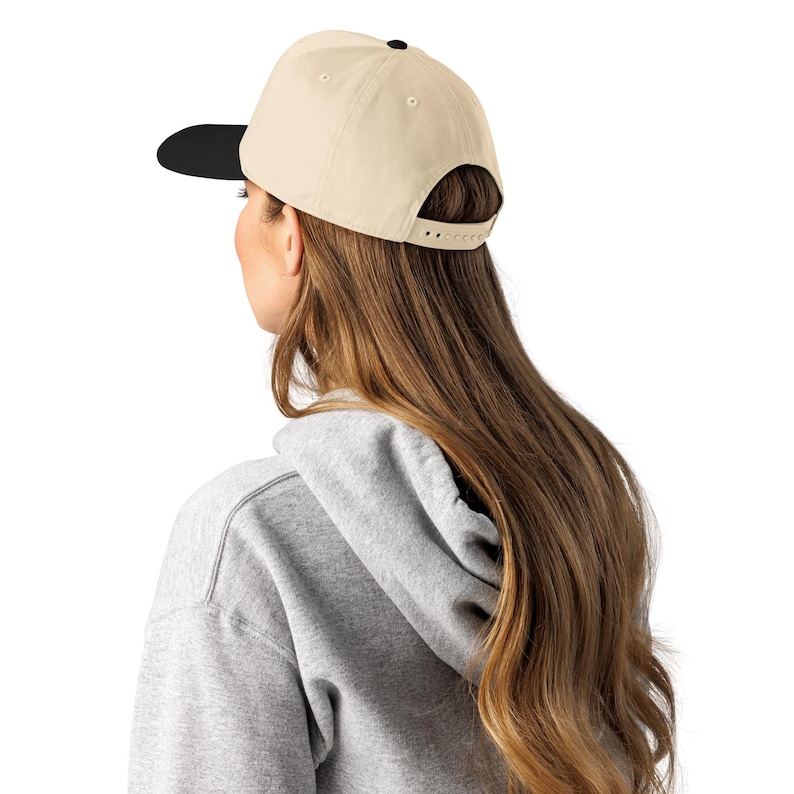 May include: A beige baseball cap with a black brim and a black button on top. The cap is worn by a person with long brown hair and a gray hoodie.