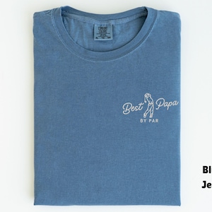 Op de afbeelding: Een blauw spijkerbroekkleurig t-shirt met de woorden "Best Papa by Par" in witte letters. Het shirt heeft een ronde hals en een klein label in het midden. Het shirt is netjes op een wit oppervlak gevouwen.