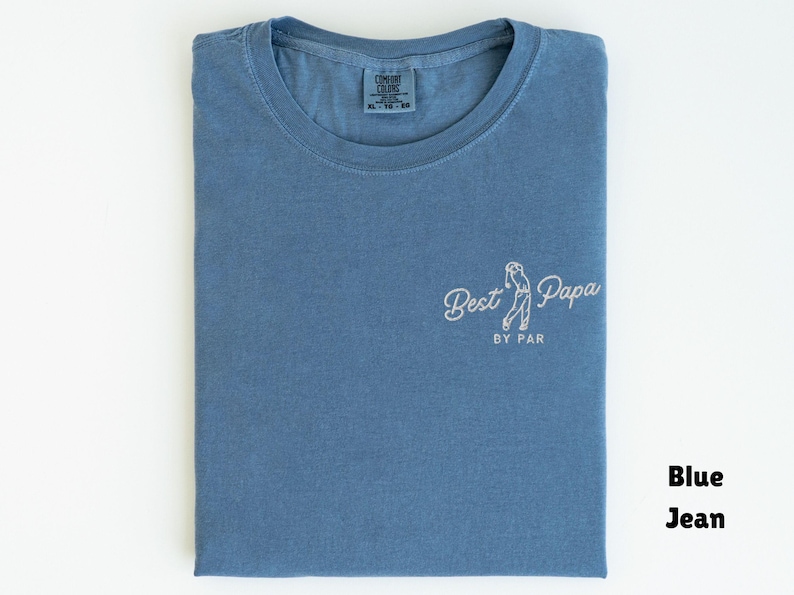 Op de afbeelding: Een blauw spijkerbroekkleurig t-shirt met de woorden "Best Papa by Par" in witte letters. Het shirt heeft een ronde hals en een klein label in het midden. Het shirt is netjes op een wit oppervlak gevouwen.