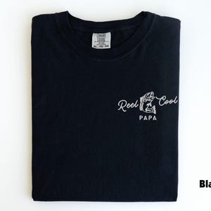Op de afbeelding: Zwart T-shirt met witte tekst "Reel Cool PAPA" en een visafbeelding. Het shirt is op een witte ondergrond gevouwen. Het label luidt XL-TG-60.