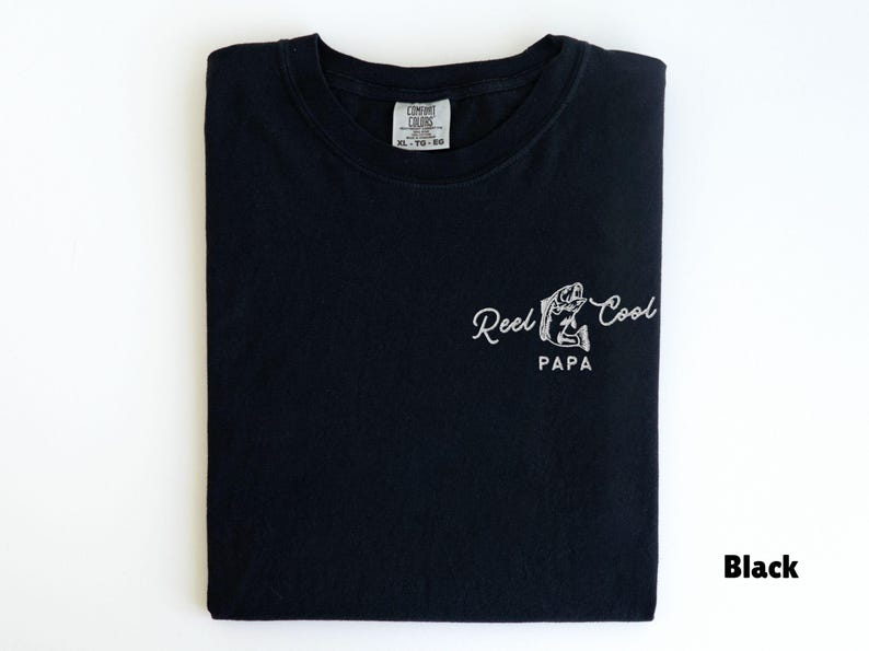Op de afbeelding: Zwart T-shirt met witte tekst "Reel Cool PAPA" en een visafbeelding. Het shirt is op een witte ondergrond gevouwen. Het label luidt XL-TG-60.