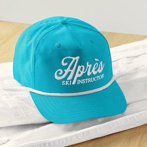 May include: A turquoise baseball cap with the word "Après" embroidered in white script above "SKI INSTRUCTOR". The cap has a white rope detail along the brim and is set against a white background.