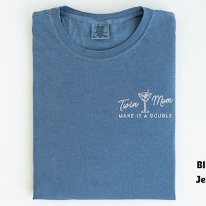 May include: A blue jean-colored t-shirt with the words "Twin Mom" and a cocktail glass graphic, along with the phrase "Make it a Double" in white script. The shirt is folded neatly.