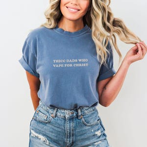 Puede incluir: Una camiseta azul con las mangas dobladas. La camiseta tiene el texto "THICC DADS WHO VAPE FOR CHRIST" en color claro. La persona lleva pantalones cortos vaqueros.
