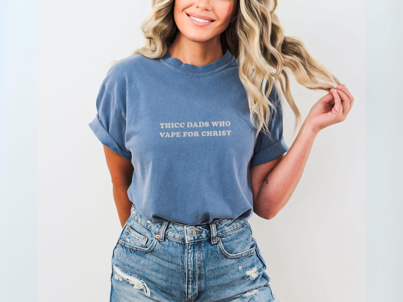 Puede incluir: Una camiseta azul con las mangas dobladas. La camiseta tiene el texto "THICC DADS WHO VAPE FOR CHRIST" en color claro. La persona lleva pantalones cortos vaqueros.