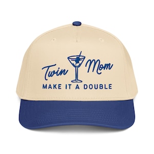 May include: A beige and blue baseball cap with the text "Twin Mom MAKE IT A DOUBLE" in blue script. The cap features a blue cocktail glass graphic. The brim and button are blue.