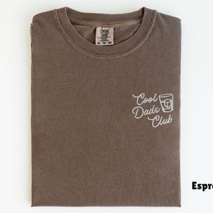 May include: Espresso-colored t-shirt with the embroidered text "Cool Dads Club" and a graphic of a cocktail glass. The shirt is folded and has a tag in the upper center. The shirt is a size XL-TG-XXG.