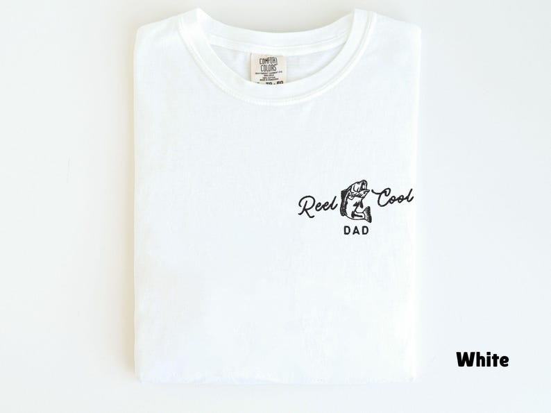 Pu&ograve; includere: T-shirt bianca con scollo tondo, piegata con cura. La maglietta presenta una grafica nera di un pesce e le parole "Reel Cool Dad" in un carattere corsivo. La maglietta &egrave; realizzata in un materiale morbido.