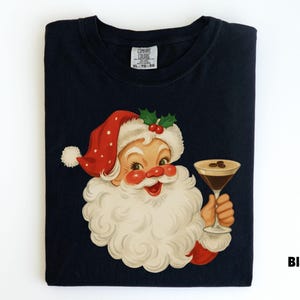 May include: Black t-shirt featuring a vintage illustration of Santa Claus holding a martini glass. Santa is depicted with a red hat with white polka dots, a white beard, and a jolly expression. The shirt has a tag that says "Comfort Colors".