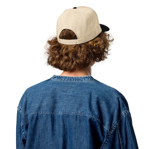 May include: A beige baseball cap with a black brim is worn backward. The cap has a flat brim and a snapback closure. The person is wearing a denim shirt.
