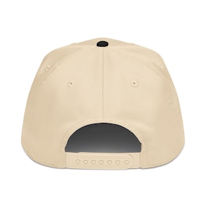 May include: A beige baseball cap with a curved peak, a black button on top, and an adjustable snapback closure. The cap has six ventilation eyelets and a structured crown. The back of the cap is visible.