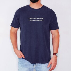 Puede incluir: Camiseta azul marino con el texto blanco "THICC DADS WHO VAPE FOR CHRIST". La camiseta la lleva una persona con vaqueros azules y un reloj.