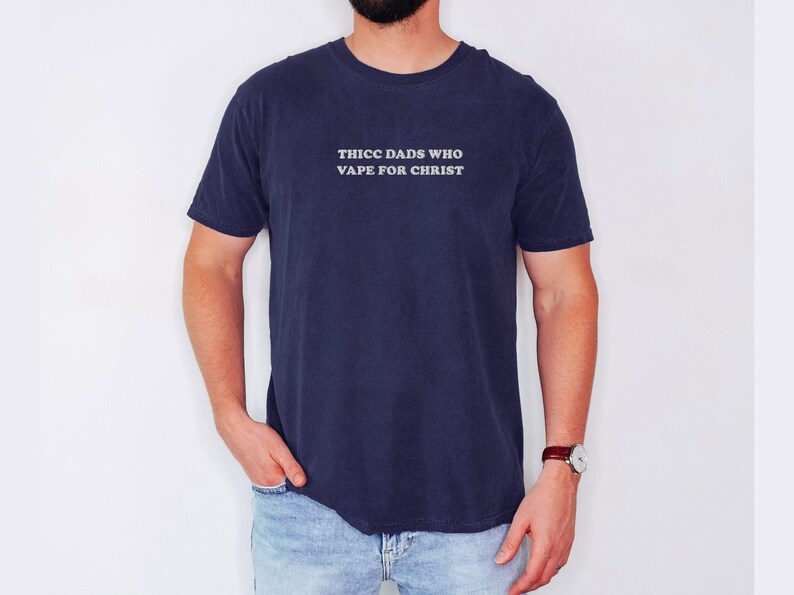 Puede incluir: Camiseta azul marino con el texto blanco "THICC DADS WHO VAPE FOR CHRIST". La camiseta la lleva una persona con vaqueros azules y un reloj.