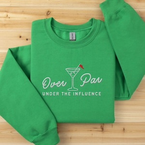 May include: A green sweatshirt with the embroidered phrase "Over Par Under the Influence" and a martini glass with a red flag. The sweatshirt is made of a soft material and is displayed on a wooden surface.