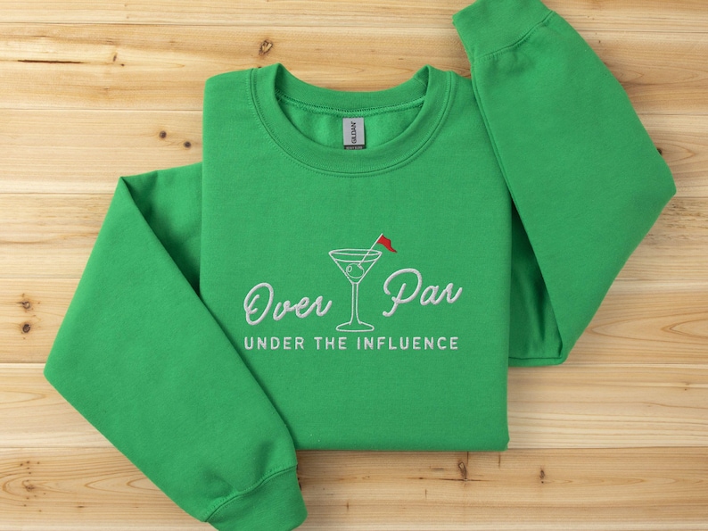 May include: A green sweatshirt with the embroidered phrase "Over Par Under the Influence" and a martini glass with a red flag. The sweatshirt is made of a soft material and is displayed on a wooden surface.