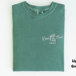 Op de afbeelding: Lichtgroen T-shirt met de tekst "Reel Cool Papa" in een sierlijk lettertype, vergezeld van een visafbeelding. Het shirt is netjes opgevouwen en toont het ontwerp op de voorkant. Het shirt heeft een effen lichtgroene kleur.
