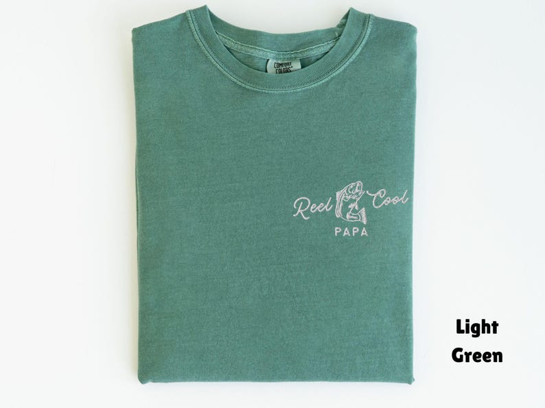 Op de afbeelding: Lichtgroen T-shirt met de tekst "Reel Cool Papa" in een sierlijk lettertype, vergezeld van een visafbeelding. Het shirt is netjes opgevouwen en toont het ontwerp op de voorkant. Het shirt heeft een effen lichtgroene kleur.