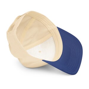 May include: A baseball cap with a cream-colored crown and a navy blue brim. The interior of the cap is cream-colored with a white lining. The cap has an adjustable snapback closure.