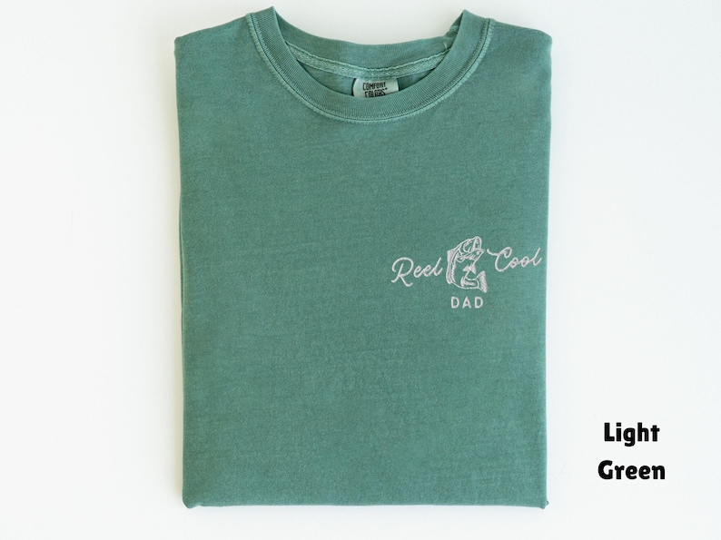 Pu&ograve; includere: T-shirt verde chiaro con la scritta "Reel Cool Dad" e una grafica di pesce. La maglietta &egrave; piegata e appoggiata su una superficie bianca. Il testo "Light Green" &egrave; nell'angolo in basso a destra.