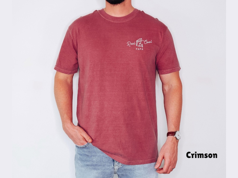 Op de afbeelding: Karmizijnrood T-shirt met de tekst "Reel Cool Papa" in witte letters. Het shirt met korte mouwen heeft een ronde hals. Het model draagt een blauwe spijkerbroek en een horloge met een bruine leren band.
