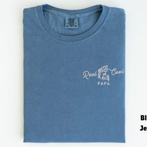 Op de afbeelding: Blauw spijkerbroekkleurig T-shirt met de tekst "Reel Cool Papa" en een visafbeelding. Het shirt is netjes op een wit oppervlak gevouwen. Het shirt is maat XL.