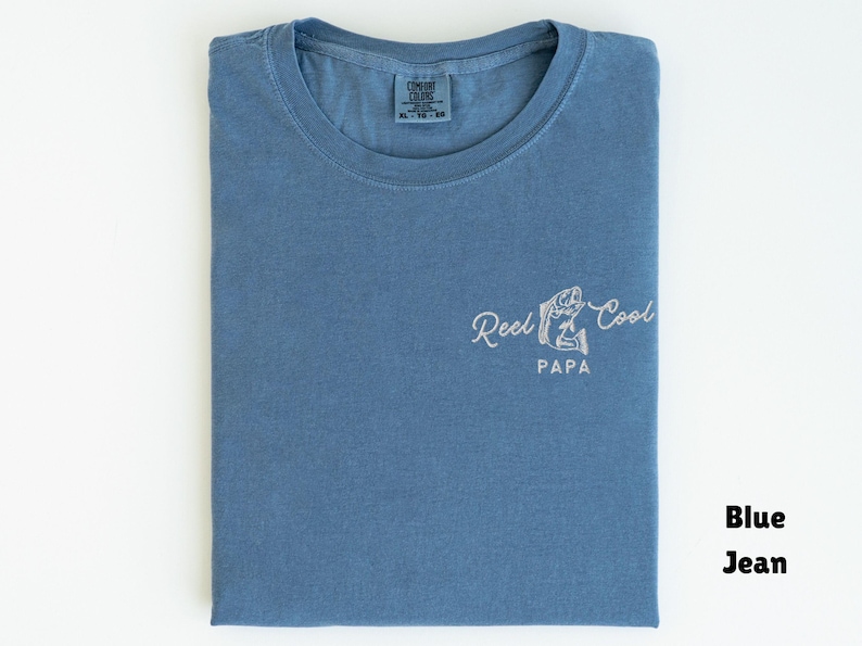 Op de afbeelding: Blauw spijkerbroekkleurig T-shirt met de tekst "Reel Cool Papa" en een visafbeelding. Het shirt is netjes op een wit oppervlak gevouwen. Het shirt is maat XL.