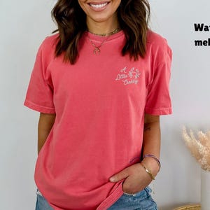 May include: A coral-colored t-shirt with short sleeves and the text "A Little Crabby" with a crab graphic. The shirt is a solid color and has a relaxed fit. The shirt is labeled "Watermelon".