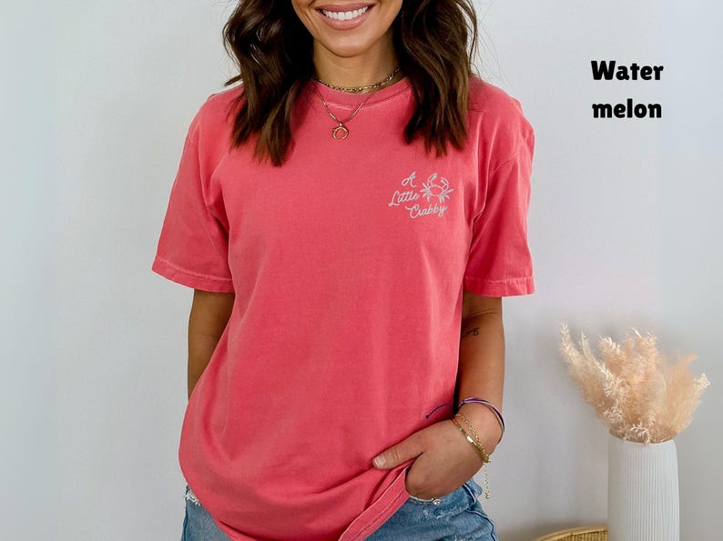 May include: A coral-colored t-shirt with short sleeves and the text "A Little Crabby" with a crab graphic. The shirt is a solid color and has a relaxed fit. The shirt is labeled "Watermelon".