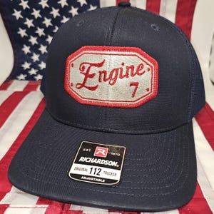 May include: A navy blue trucker hat with a red and white patch that says "Engine 7". The hat is adjustable and has a Richardson 112 label.