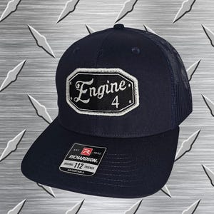 May include: A navy blue trucker hat with a mesh back and a flat brim. The front features a black octagonal patch with white embroidered trim and the white text Engine 4. A black and white sticker on the brim reads Richardson 112 Trucker Adjustable.