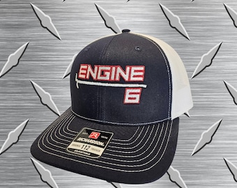 May include: A navy blue and white trucker hat featuring an embroidered red and white patch that reads ENGINE 6 with a horizontal line graphic. The hat has a structured front panel, a mesh back, and a black brim with a Richardson 112 sticker attached.