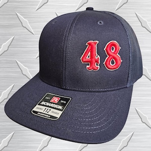 May include: A navy blue trucker hat featuring the number 48 embroidered in red with a white outline on the front panel. The hat includes a black sticker on the brim with the text Richardson 112 Trucker Adjustable Est 1970 and a logo.