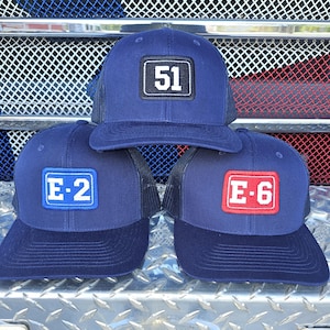 May include: Three navy blue trucker hats with black mesh back panels. The hats have different embroidered patches. One patch is black with white text that says "51". Another patch is blue with white text that says "E-2". The third patch is red with white text that says "E-6".