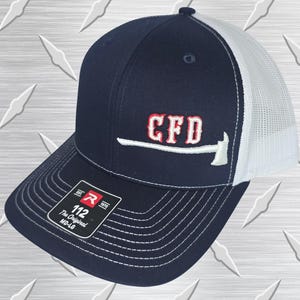 May include: A navy blue and white trucker hat featuring an embroidered red CFD logo above a white axe graphic. The front panel displays a black sticker with the text R 1970 112 The Original MD-LG. The hat has a structured crown and mesh back.
